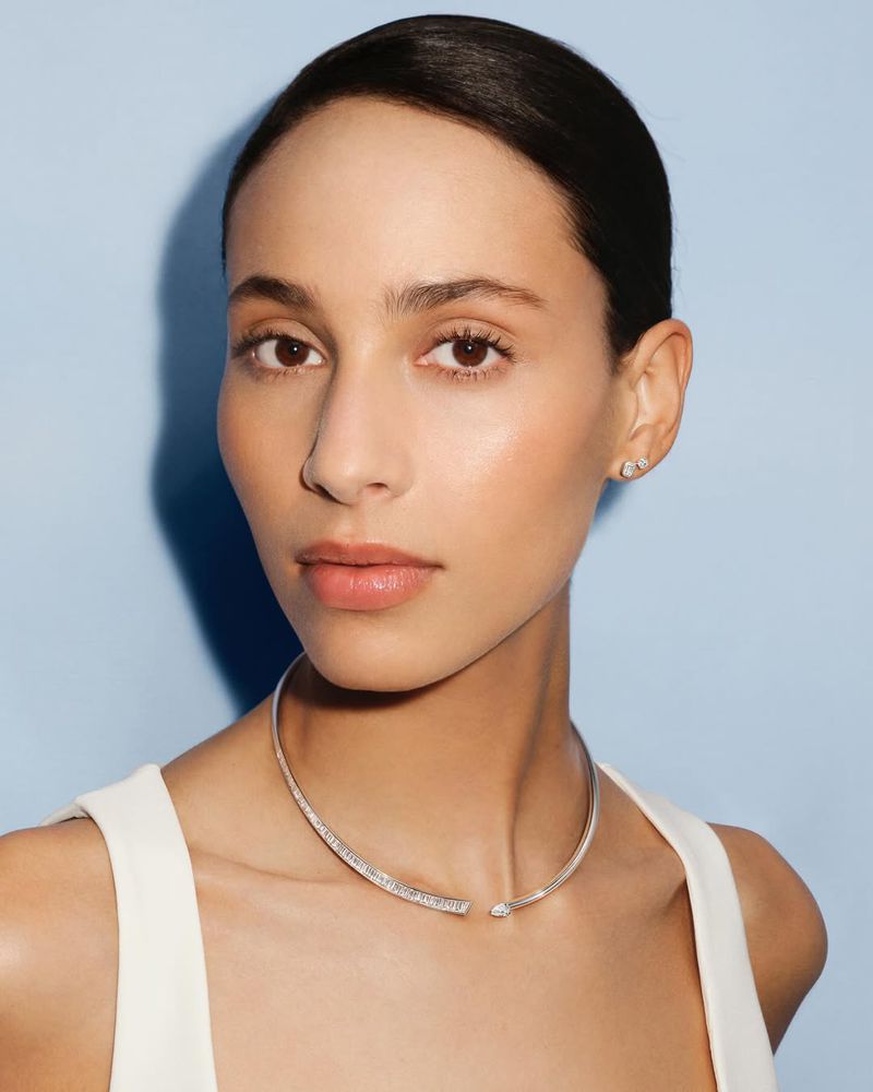 Close-up shot of a woman with fair skin wearing a white top and a silver choker necklace, posing against a light blue background.