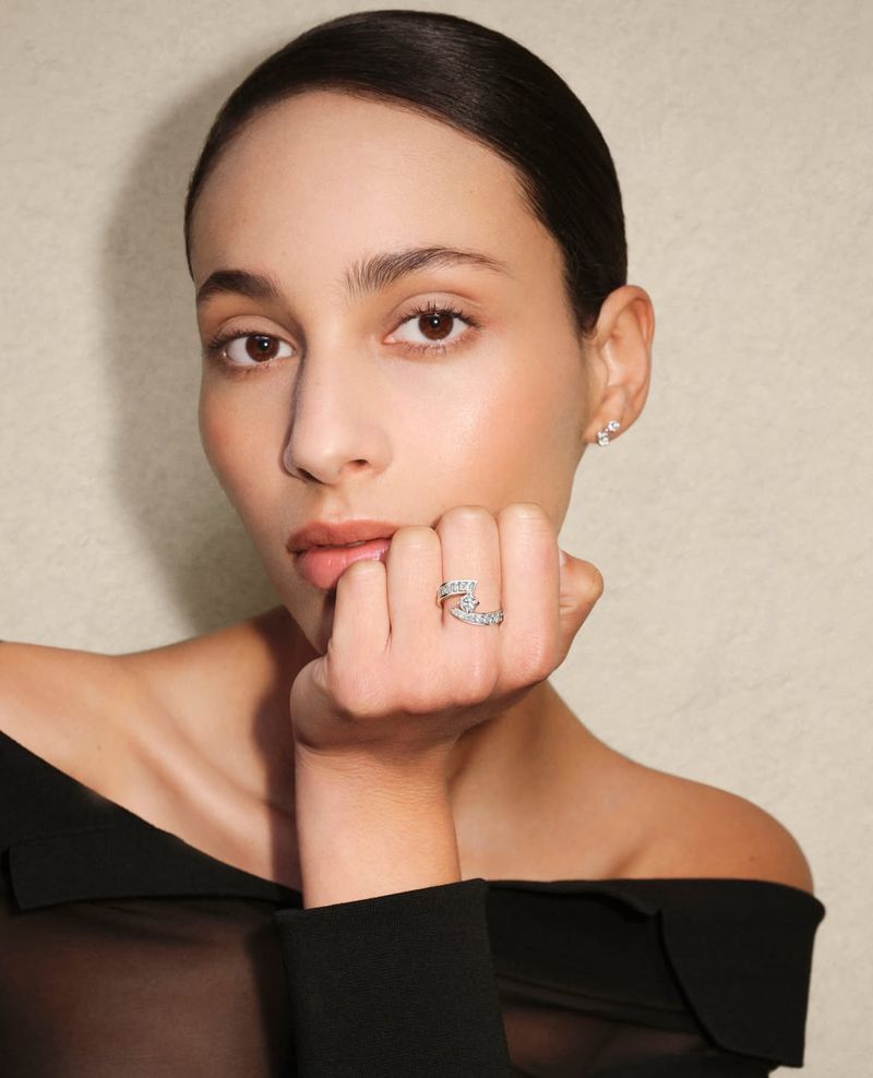 Close-up shot of a woman with her hand near her face, showcasing a ring on her finger. The background is a neutral color.