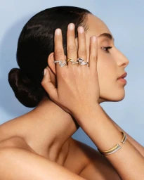 Close-up profile of a model posing with hands up displaying diamond rings and bracelets against a light blue studio background.