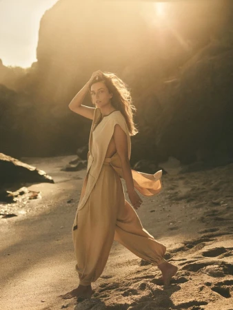 A woman in casual clothing stands on a sandy beach with rocky cliffs and bright sunlight, enjoying a peaceful moment in a natural setting.