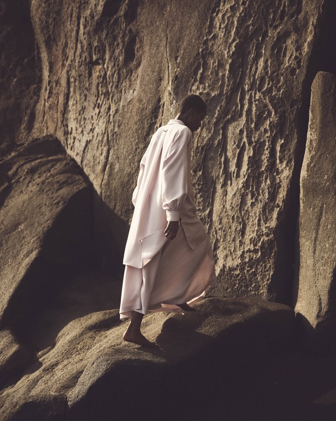 Barefoot Black male model wearing a pink outfit walking on rocks in a rugged outdoor setting with dramatic lighting and neutral tones.