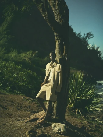 Person in light beige shorts suit poses near a tree on a grassy shore with a background of water and lush green trees under a blue sky.