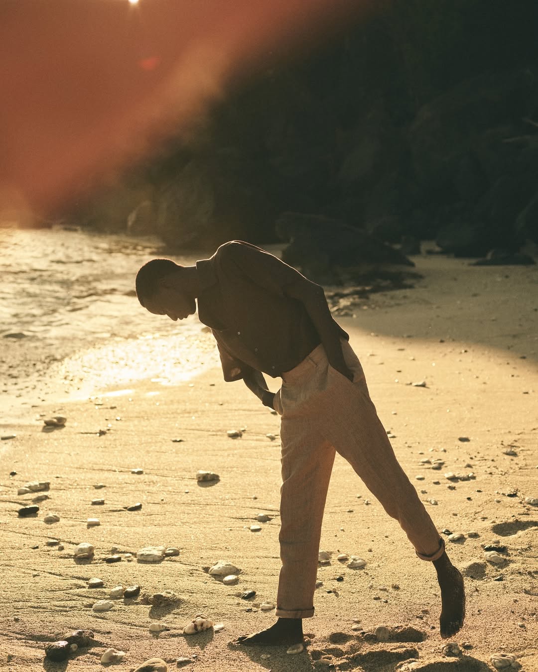 Barefoot adult male is standing on a beach with rock formations and ocean in the background, bathed in warm, golden sunlight.