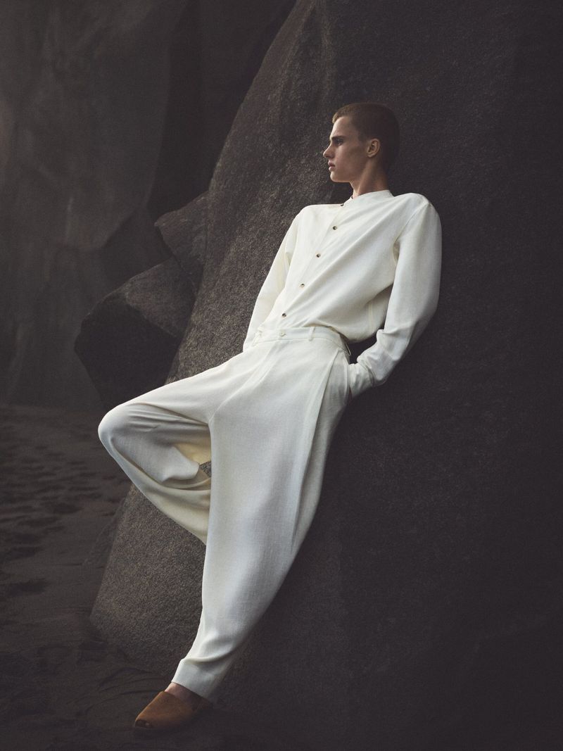 A slender adult male poses while leaning against a dark, weathered rock formation dressed in cream-colored fashion clothing and brown leather shoes.