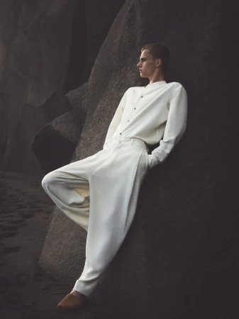 A slender adult male poses while leaning against a dark, weathered rock formation dressed in cream-colored fashion clothing and brown leather shoes.