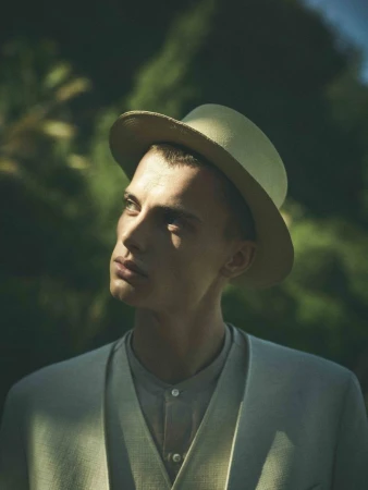 Model Elias Monstrey wearing a light beige hat and tailored suit looking upward against a blurred green foliage background.
