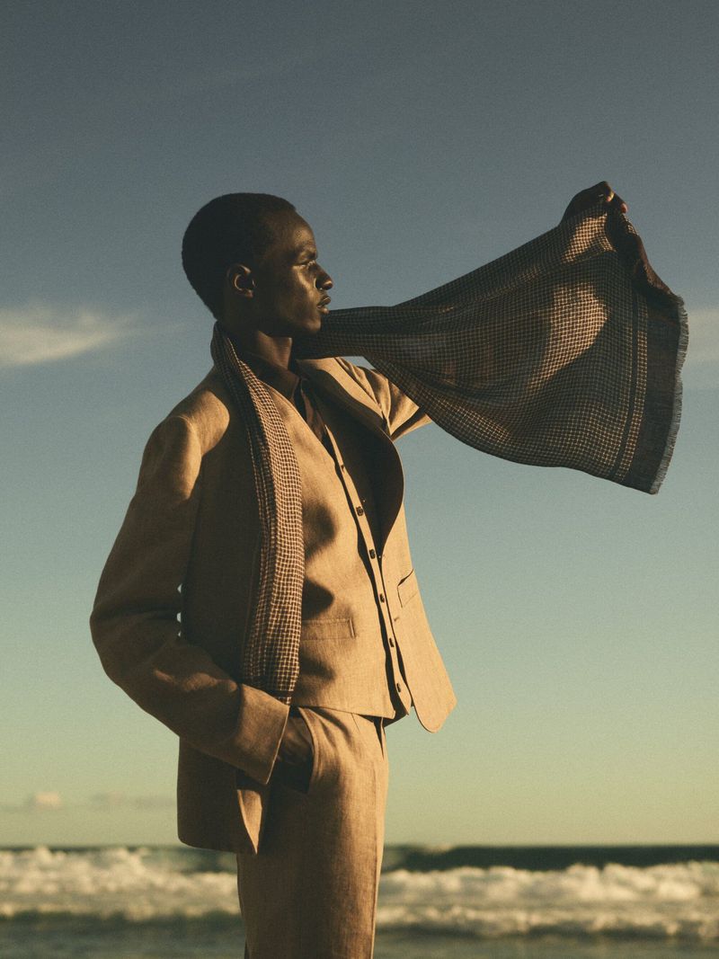 Fashionable adult in suit holding shawl, standing near wavy ocean and clear sky during daytime in outdoor editorial fashion shot.
