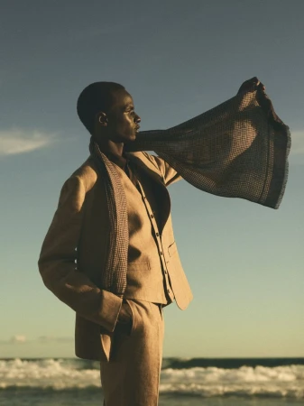 Fashionable adult in suit holding shawl, standing near wavy ocean and clear sky during daytime in outdoor editorial fashion shot.