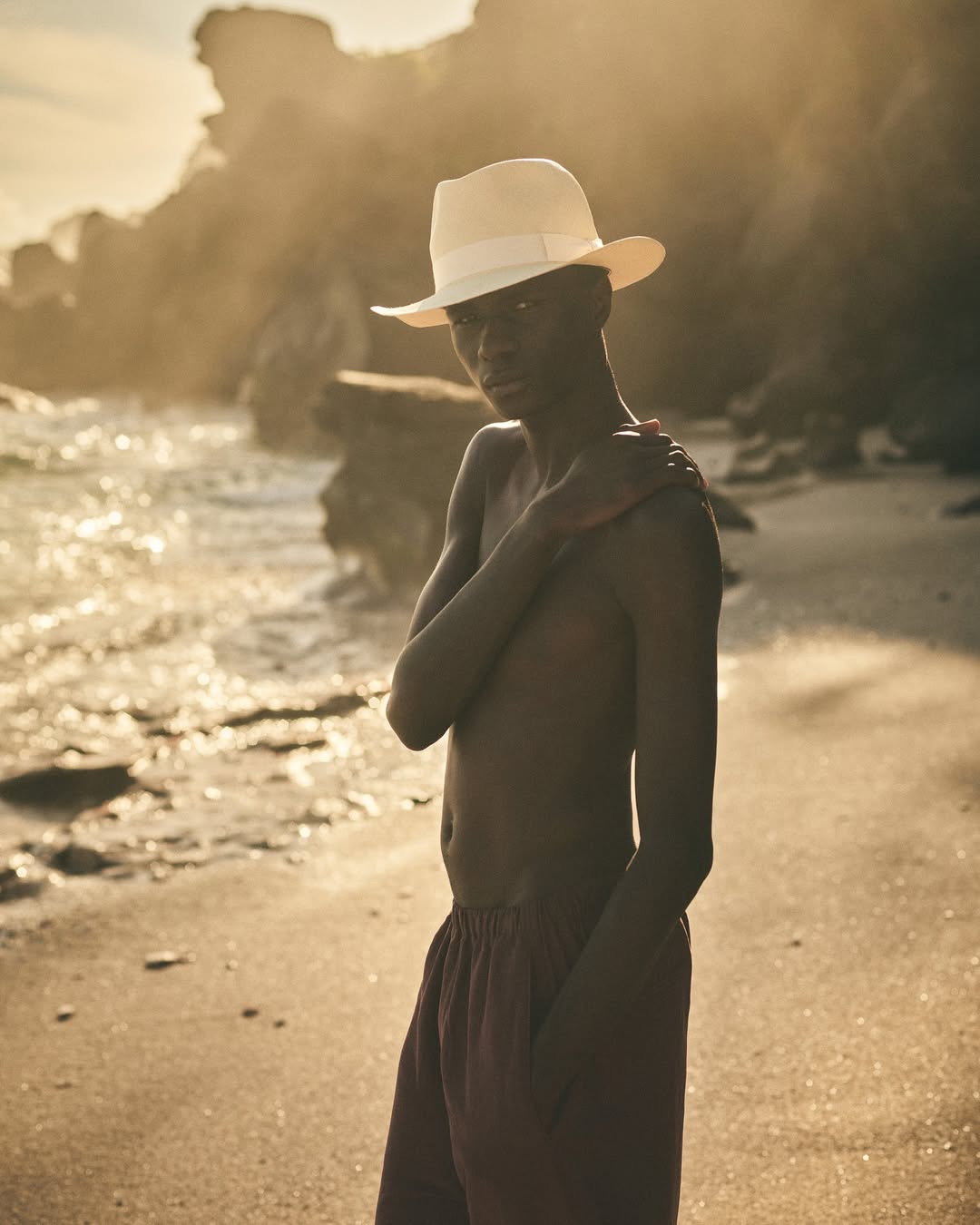 Person on beach wearing hat. Dark-skinned shirtless person in red pants. Golden sunlight, ocean background, rocky shoreline.