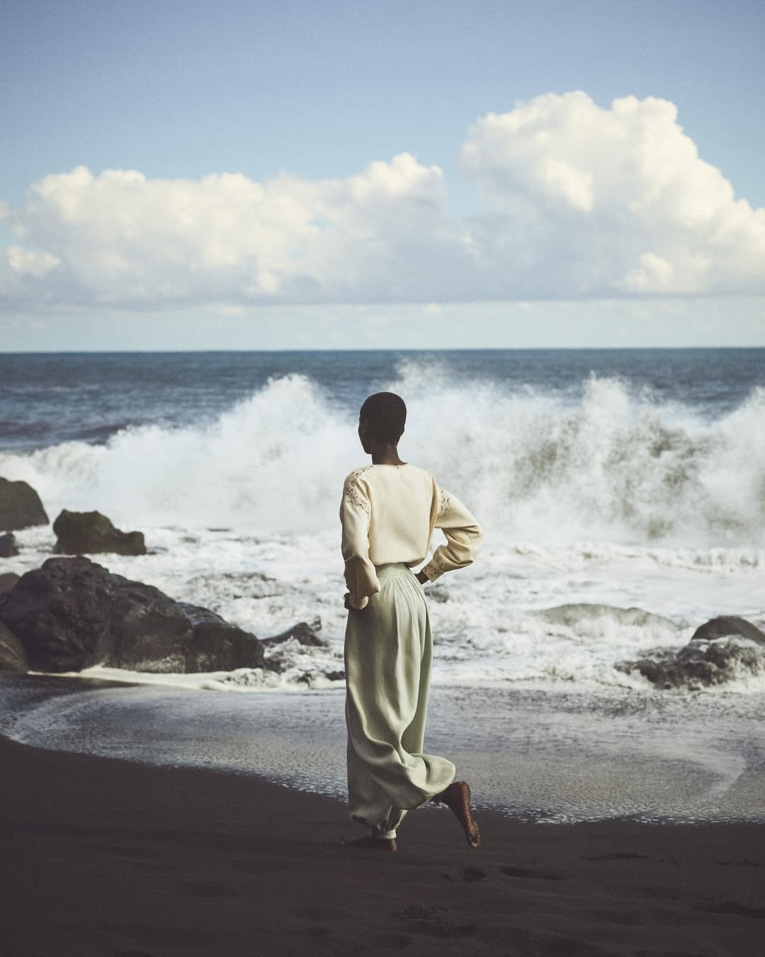 Woman in billowing pants walks on black-sand beach with crashing ocean waves, large rock formations, and cloudy blue sky.