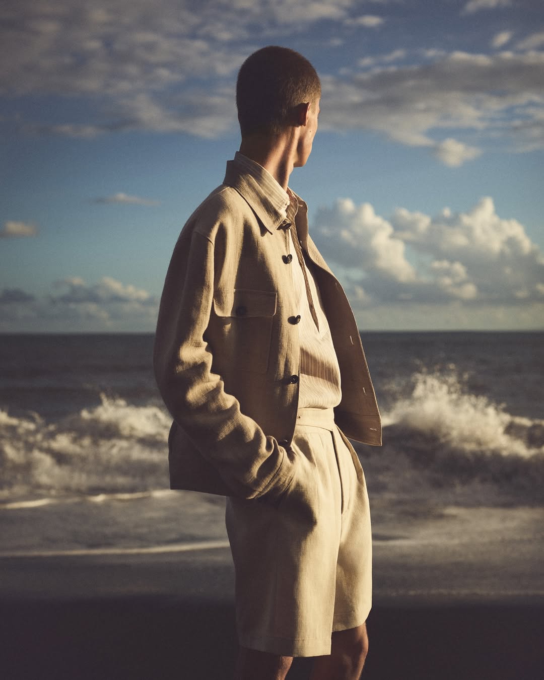 Adult male wearing a linen shirt and shorts stands on the beach with ocean and sky in the background, fashion-oriented, looking away.
