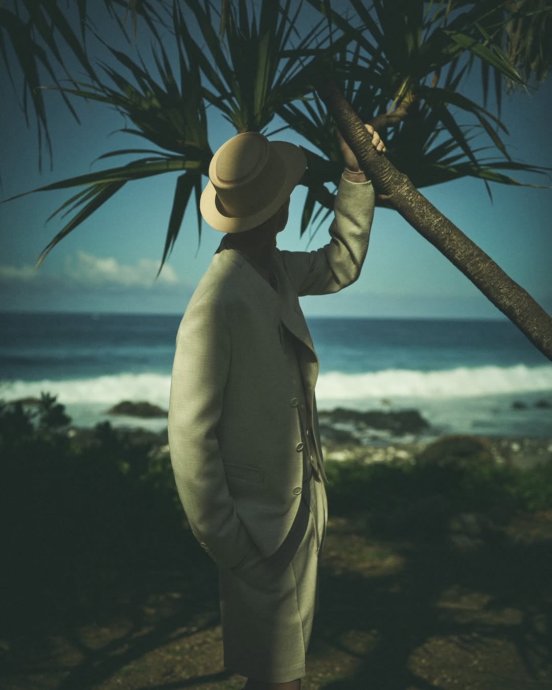 A man wearing a tailored beige suit and matching brimmed hat stands next to a palm tree overlooking the ocean.
