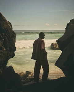 Model standing on a rocky shoreline looking out toward the ocean for Loro Piana Resort 2025 campaign.