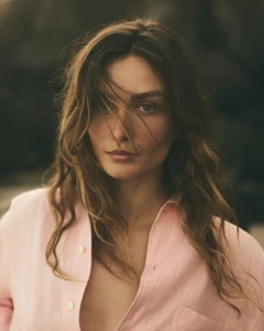 Portrait of a woman wearing a light pink button-down shirt with loose wavy hair framing her face outdoors.