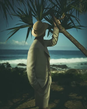 A man wearing a tailored beige suit and matching brimmed hat stands next to a palm tree overlooking the ocean.