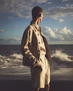 Male model in beige jacket and matching shorts standing on a sandy beach against an ocean and cloudy sky.