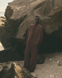 Model standing outdoors against a large rocky coastal formation wearing a dark red button shirt and matching trousers.