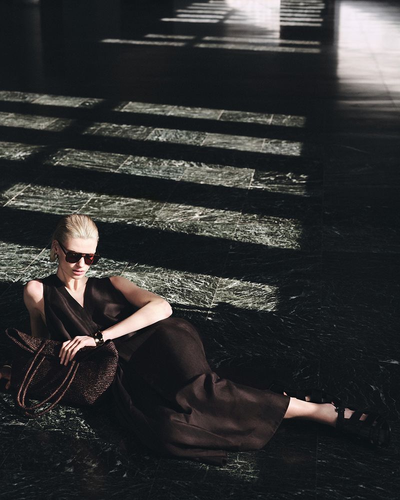 Elegant blonde model in sunglasses and a dark dress lounges on a polished, shadowed floor next to a woven handbag.