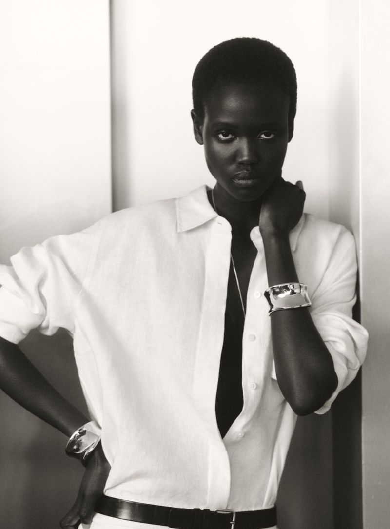 Monochrome portrait of a Black woman with short hair in a white shirt and black belt, posing against a wall with natural light.