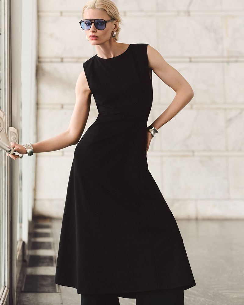 Elegant woman in a sleeveless black dress stands inside a modern marble-walled building near glass windows, showcasing fashion and style.