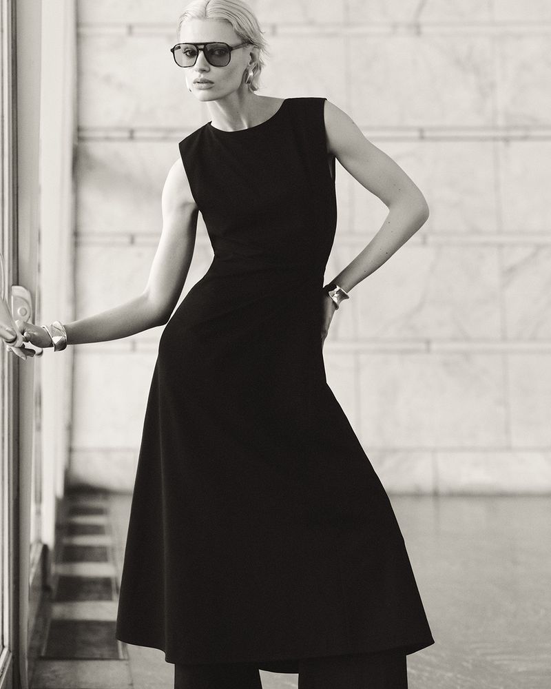 Black-and-white portrait of a woman with short blonde hair, sunglasses, and a black dress, posing in a marble setting.
