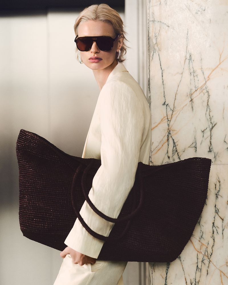 Woman in sunglasses and white suit carries an oversized brown woven tote bag against a marble wall, fashion forward and stylish.