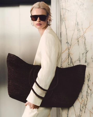 Woman in sunglasses and white suit carries an oversized brown woven tote bag against a marble wall, fashion forward and stylish.