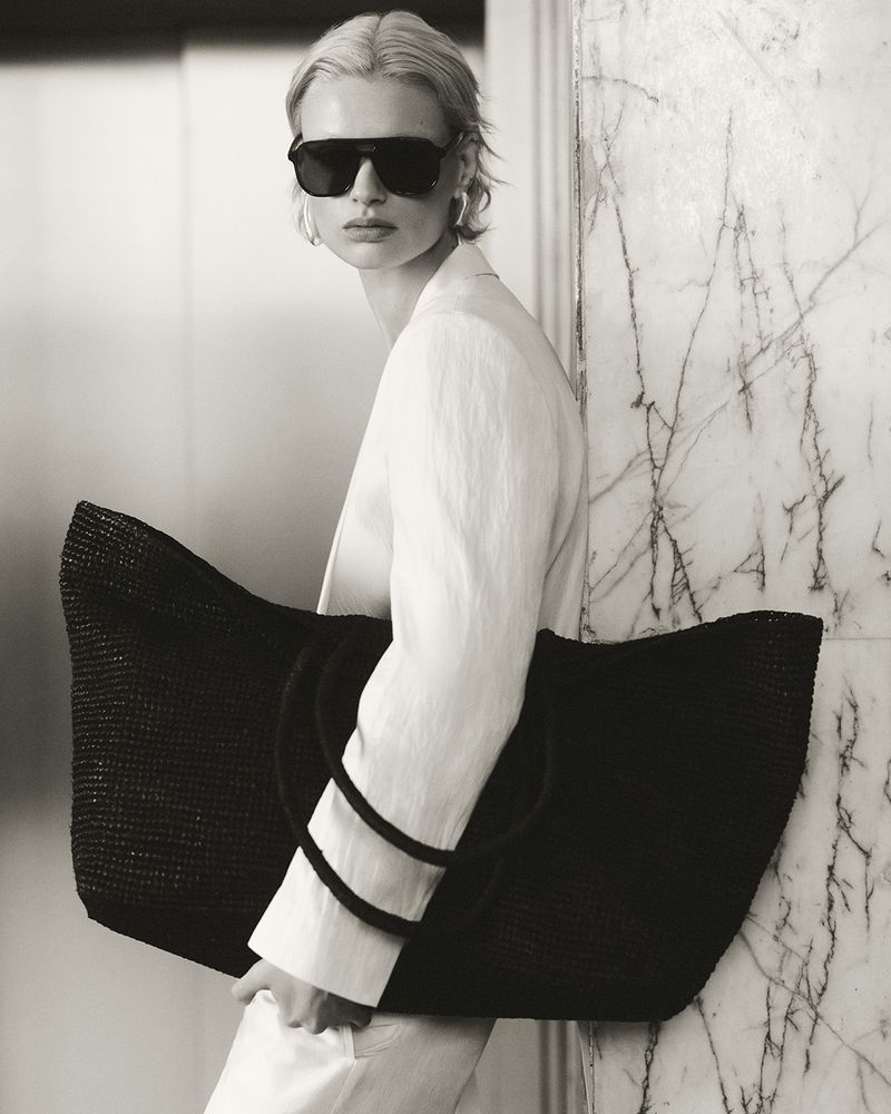 Monochrome portrait of a blonde adult woman in sunglasses wearing a white linen suit and holding a large black raffia tote bag.