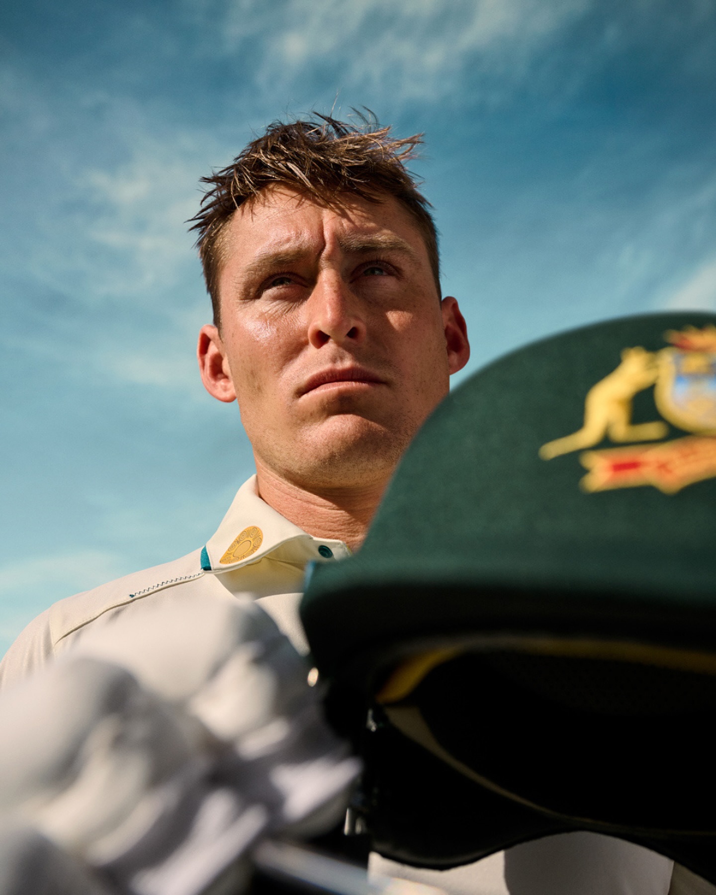 Portrait of male adult cricket player in uniform holding his green helmet with the Australian emblem, against a bright blue sky.