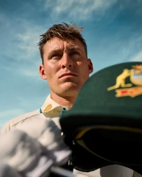 Low-angle shot of a male cricketer in whites holding a green cap against a clear blue sky, photographed by Tobias Rowles.
