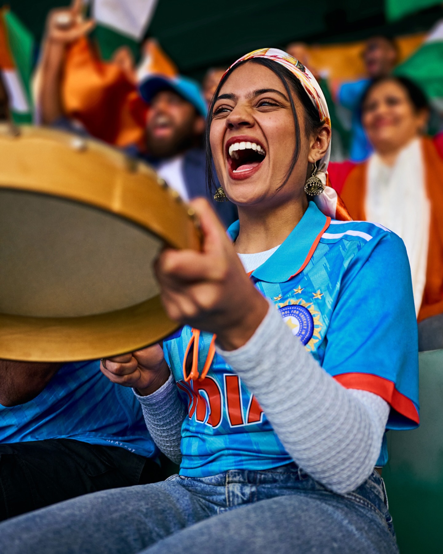 Enthusiastic cricket fan cheers loudly, wearing an India jersey and headscarf, while holding a tambourine at an outdoor game.