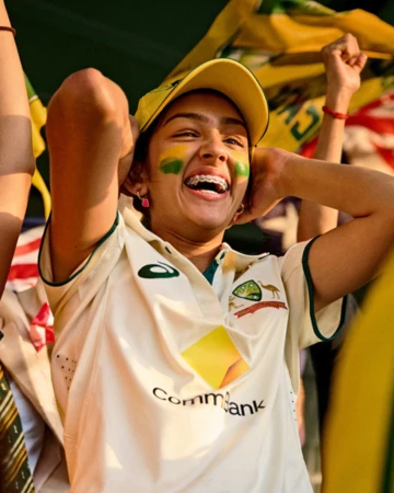 Enthusiastic young woman with face paint cheering at a cricket match, wearing team colors and cap, in a close-up, eye-level shot.