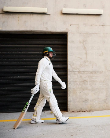 An adult male cricketer wearing a helmet and cricket whites walks along a concrete surface with a bat under his arm.