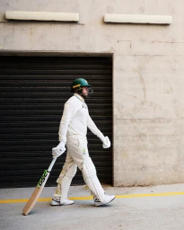 Cricket player in white uniform and green helmet walking outdoors while holding a bat
