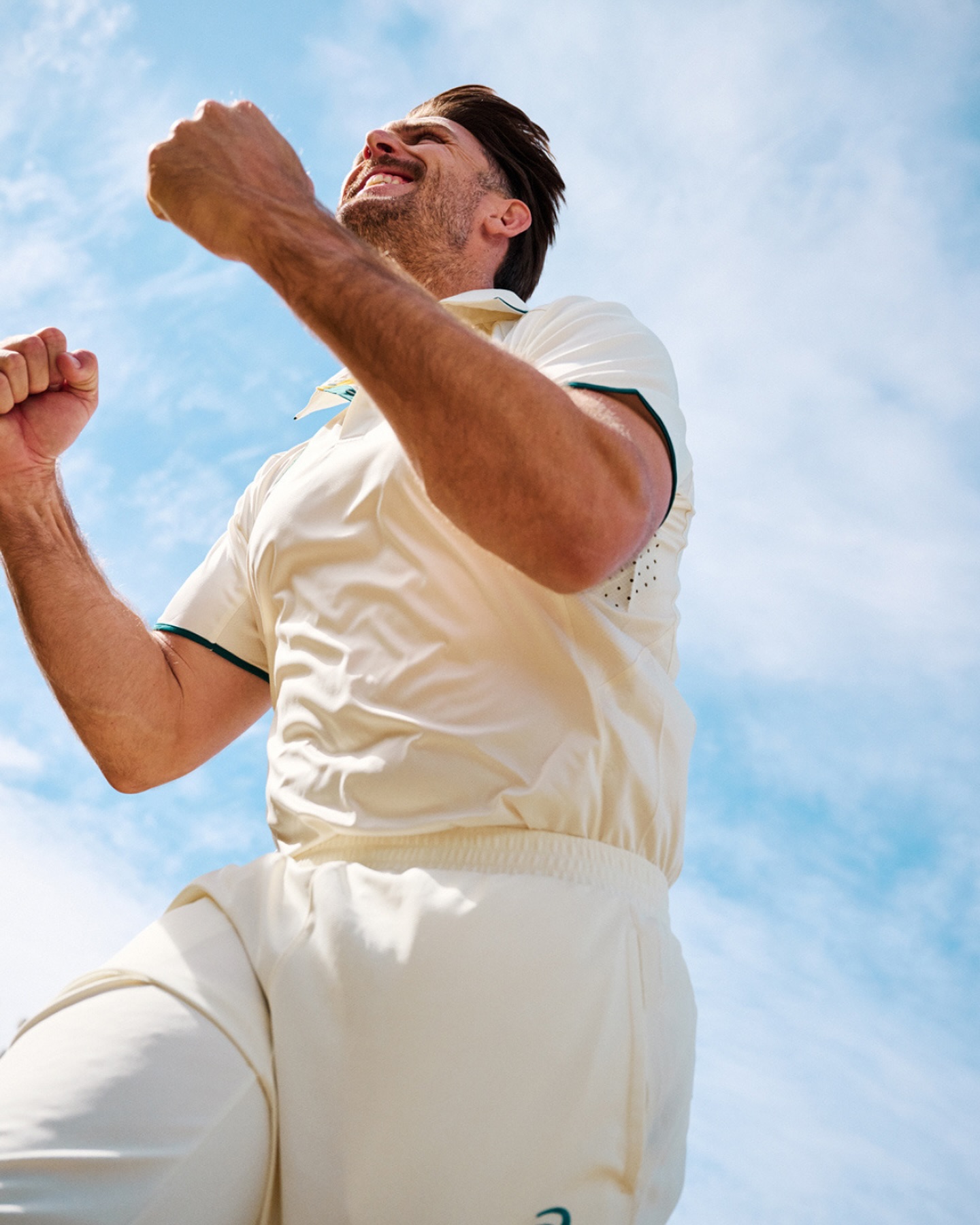 An upward shot captures a man in a cricket uniform celebrating with fists clenched, against a bright blue sky with soft, scattered clouds.