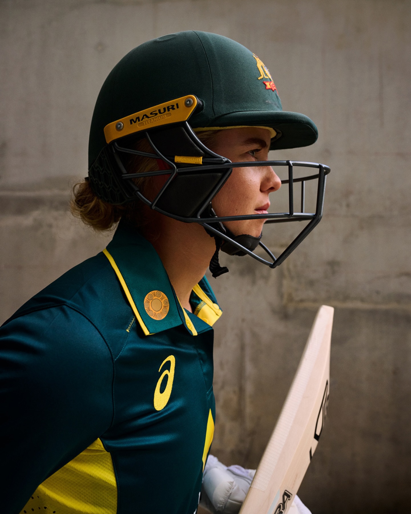 Profile portrait of a cricket player wearing a protective helmet and green and yellow jersey holding a wooden cricket bat indoors.