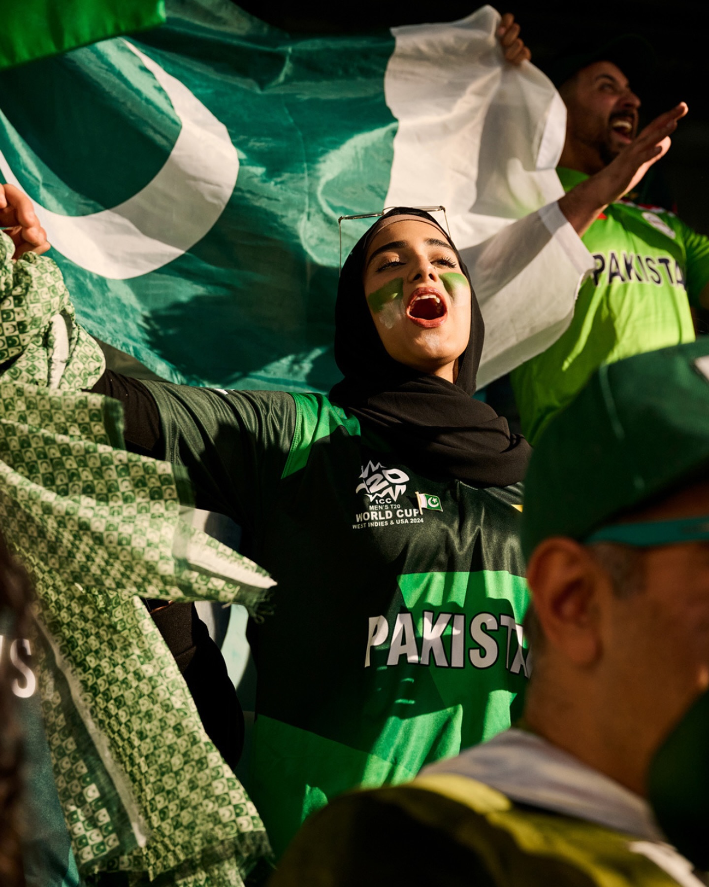 Enthusiastic cricket fan cheers with green face paint, hijab, and a Pakistan flag, showing support at an outdoor cricket match event.