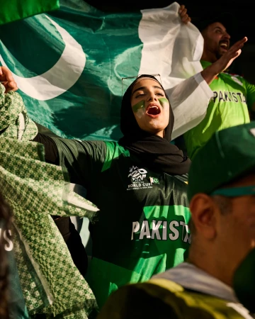 Enthusiastic cricket fan cheers with green face paint, hijab, and a Pakistan flag, showing support at an outdoor cricket match event.