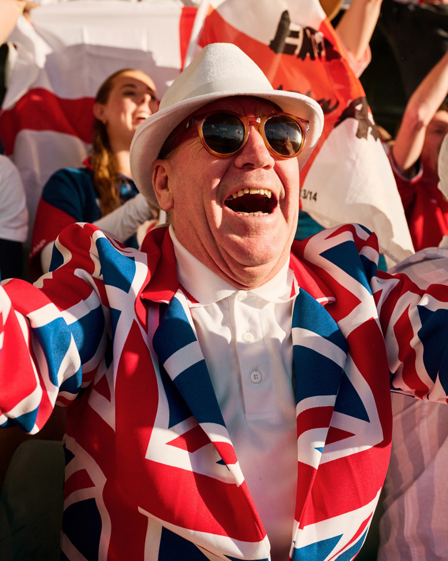 Enthusiastic mature adult man cheers wearing a Union Jack jacket and hat at an outdoor event with other fans and English flags.