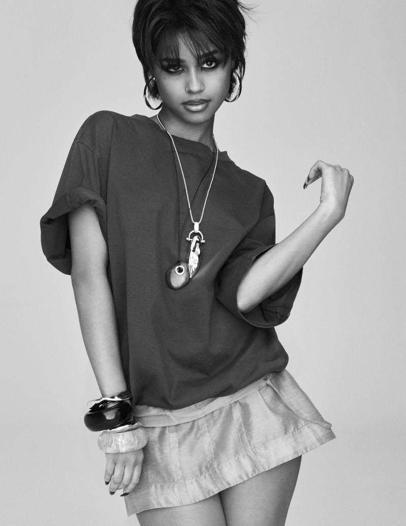 Black and white portrait of a young woman in an oversized t-shirt and mini skirt, accessorized with jewelry, posing in a photo studio.