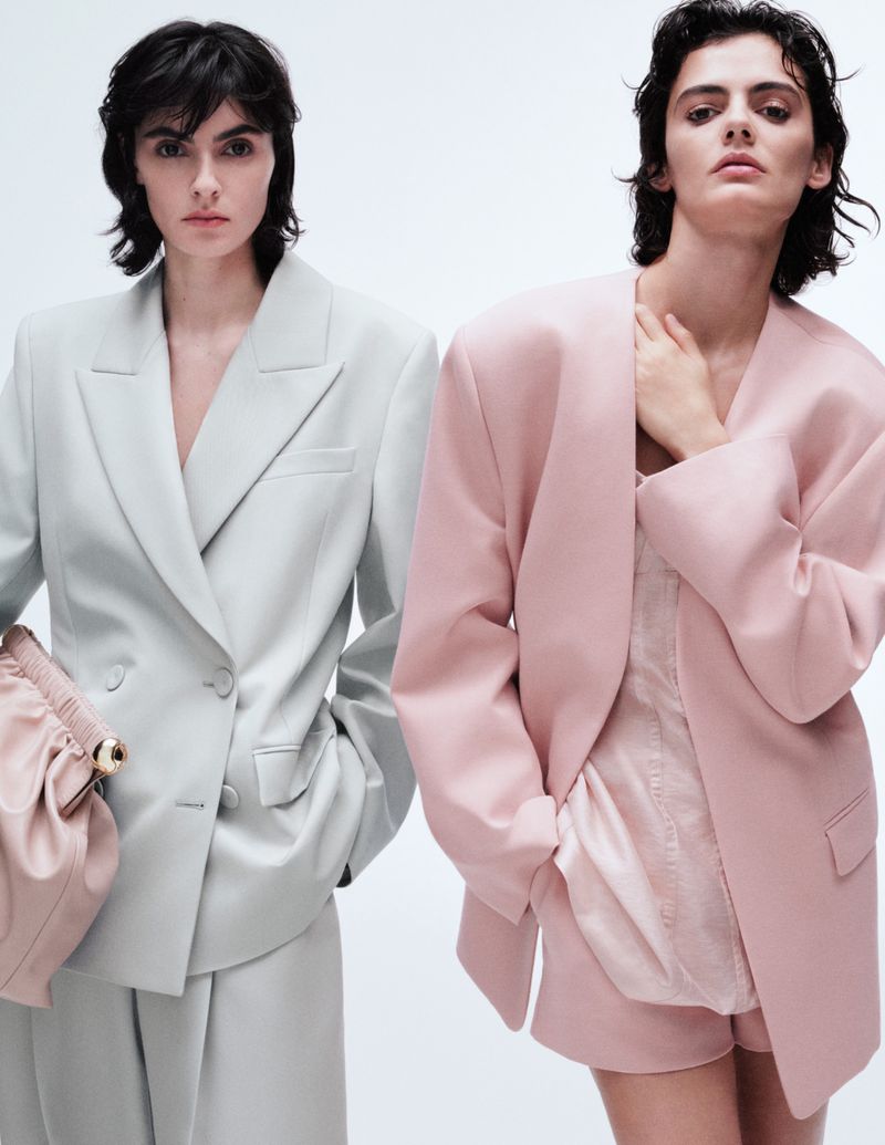 Two women pose in pastel blazers and shorts against a white backdrop, one in pale green with a matching handbag, the other in pink.