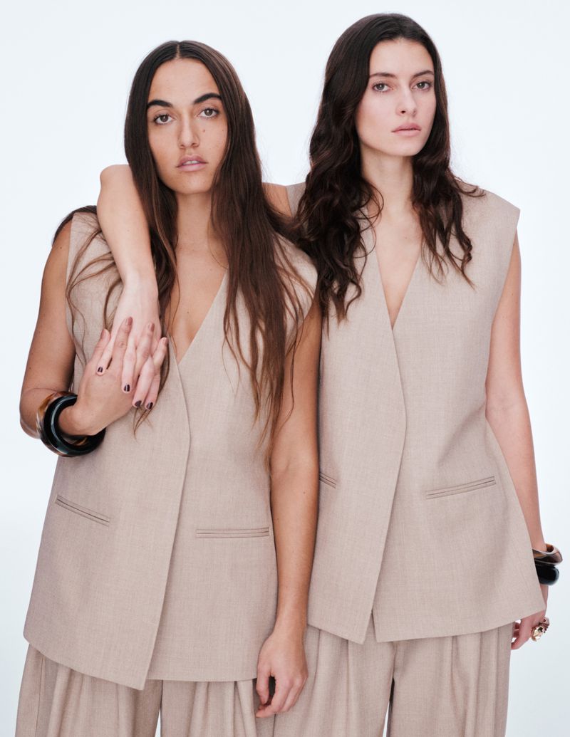 Two women pose in matching beige vest and pants outfits against a white backdrop, showcasing minimalist fashion and neutral tones.