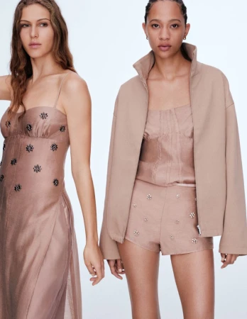 Two women pose indoors wearing fashionable light-brown outfits. The woman on the left wears a long dress, the other wears a jacket and shorts.