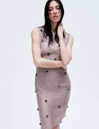 Caroline Polachek wearing a sleeveless taupe dress with scattered dark embellishments against a white background.