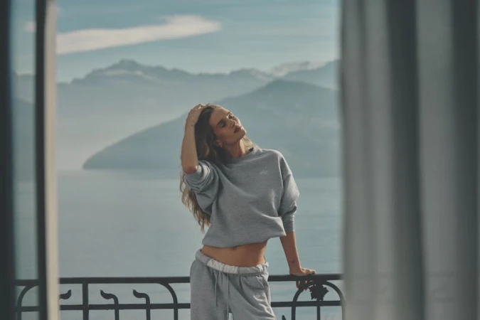 A blonde woman in grey loungewear poses on a balcony overlooking a lake and mountains, bathed in soft daylight. Fashion or travel content.