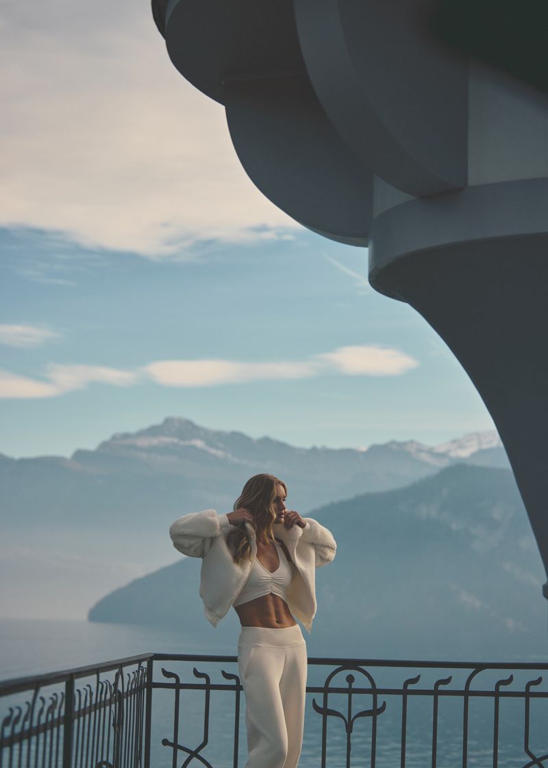 Woman in white outfit poses on a balcony with lake and mountain views in background, under a partially visible building feature.