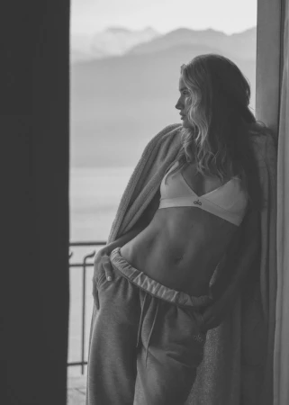 A woman with blonde hair poses in athletic wear near a window with a view of mountains and water in the background in a grayscale photograph.