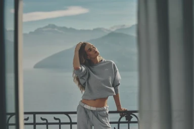 Rosie Huntington-Whiteley in a grey Alo sweatshirt and sweatpants on a balcony overlooking a lake and mountains.