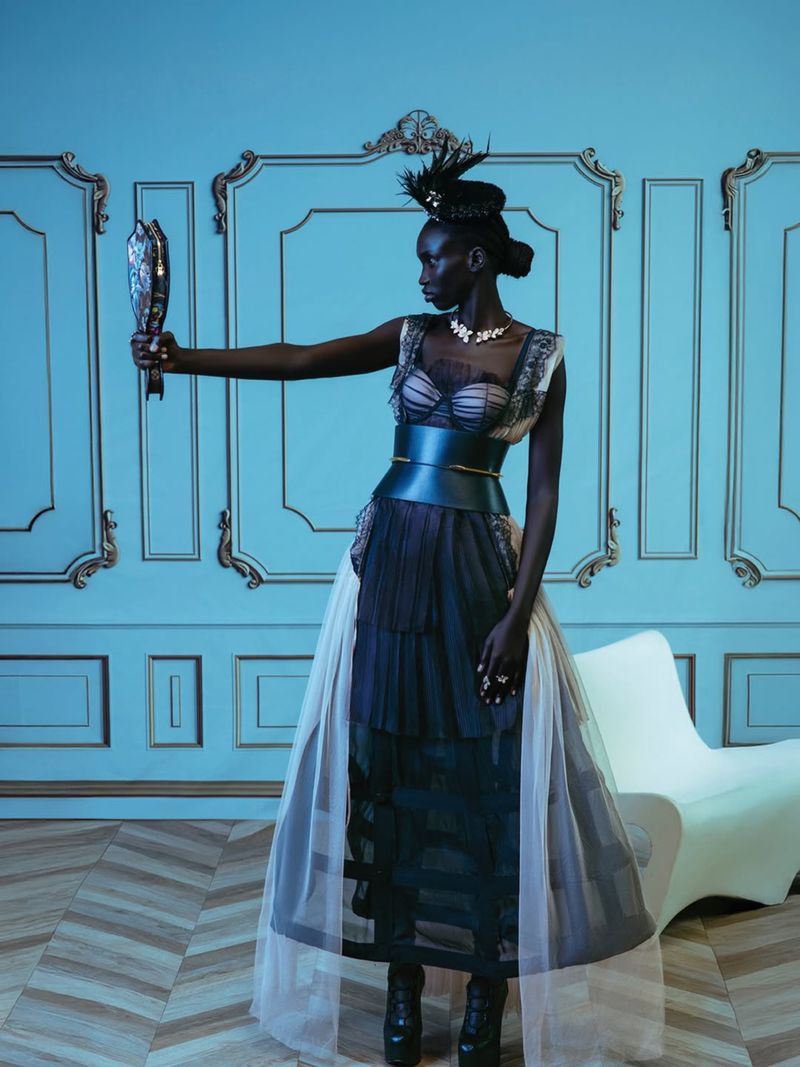 Elegant dark-skinned woman in an elaborate dress holds a mirror in an ornate, light-blue studio, showcasing a fashion-forward, artistic aesthetic.