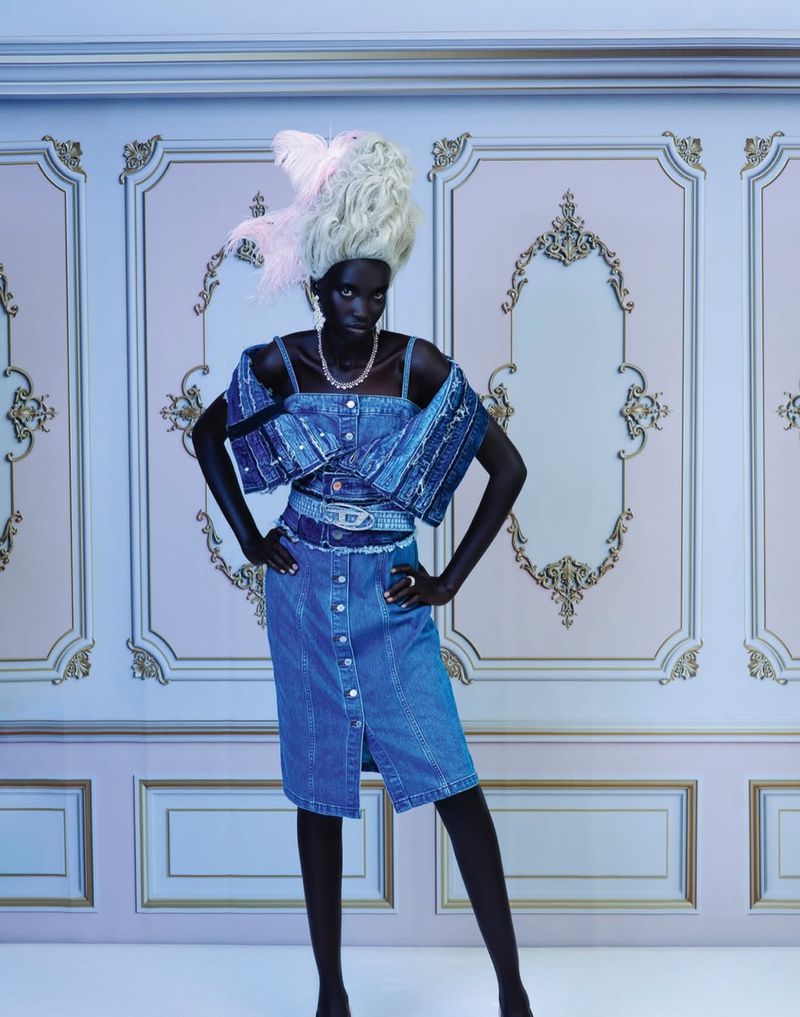 Fashion shot of a dark-skinned model wearing a denim dress and a decorative wig, posing in front of an ornate wall with molding.